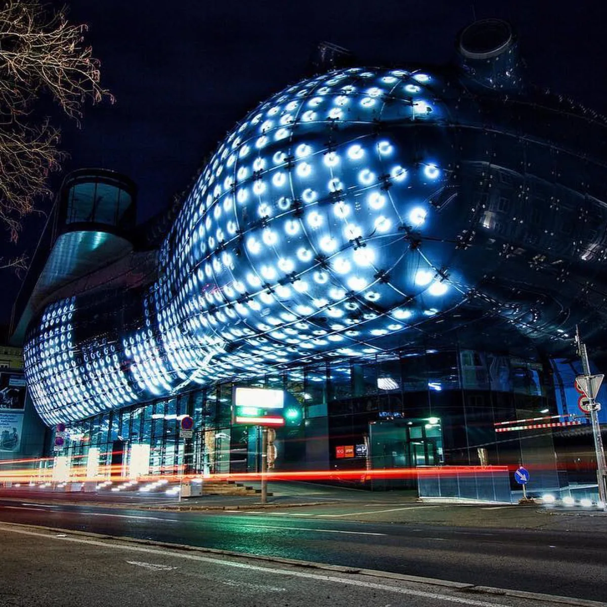 Foto malam Kunsthaus Graz di Austria, sebuah museum seni kontemporer dengan bentuk organik yang unik dan dilapisi panel LED biru yang menyal