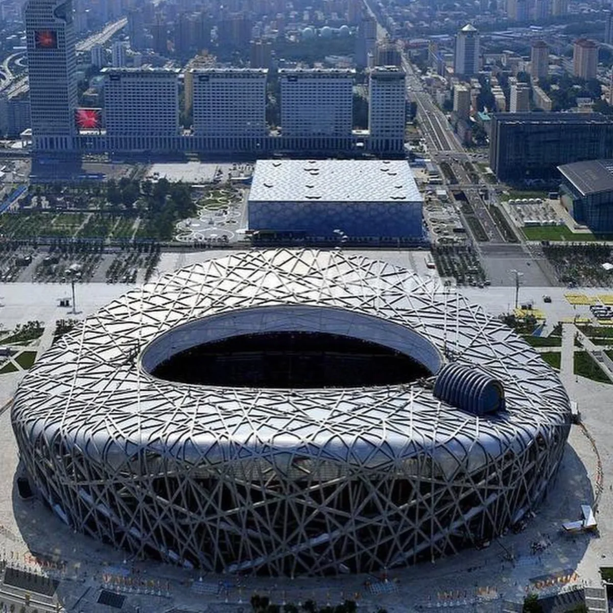 Foto udara sudut lebar Stadion Nasional Beijing (Sarang Burung) yang melingkar dan luas, menampilkan kisi-kisi anyaman balok logam yang rumit dan kacau di fasadnya, dikelilingi oleh alun-alun Olimpiade yang beraspal.