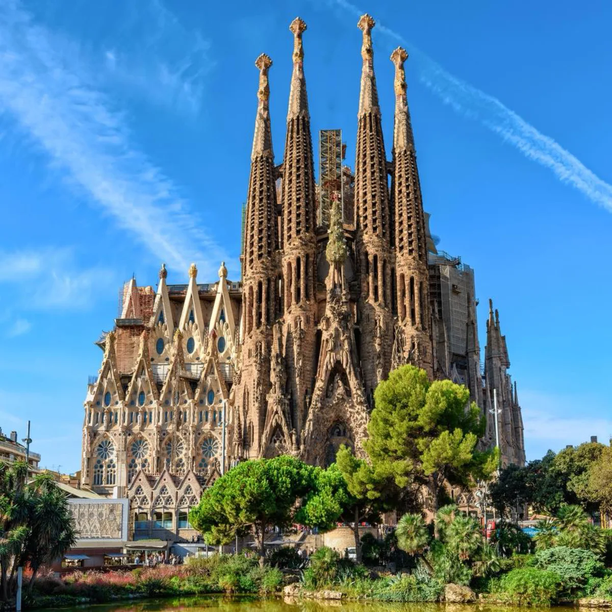 Pemandangan dari tingkat permukaan melintasi kolam taman reflektif dari fasad Nativity yang rumit dan menara lonceng utama basilika Sagrada Familia yang belum selesai di Barcelona, membubung tinggi ke langit biru cerah dengan awan, dibingkai oleh pepohonan.