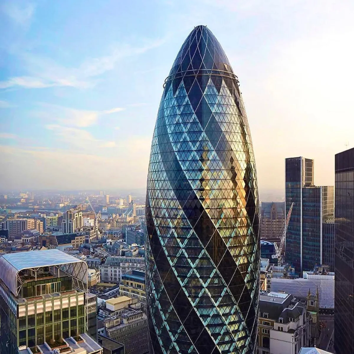 Pemandangan udara sudut tinggi dari gedung pencakar langit ikonik 30 St Mary Axe (The Gherkin) berlapis kaca, membubung tinggi di atas distrik keuangan City of London yang padat, dengan struktur diagridnya yang berputar menangkap cahaya matahari terbenam yang hangat.