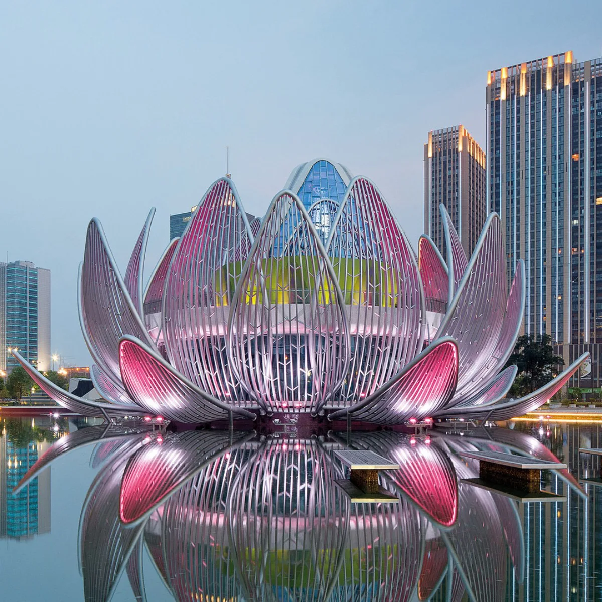 Foto senja jarak dekat dari kompleks Bangunan Teratai patung di Wujin, Tiongkok, struktur utamanya diterangi warna merah muda seperti bunga teratai mekar raksasa dengan inti yang bercahaya, menjulang dari kolam reflektif yang sempurna dengan gedung pencakar langit modern di kejauhan.