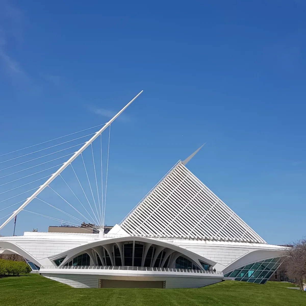 Foto tingkat permukaan dari sayap peneduh matahari putih (Brise Soleil) paviliun Museum Seni Milwaukee di Milwaukee, terbuka lebar dan menjulang ke langit biru cerah, menampilkan struktur patung modern di atas halaman rumput hijau.