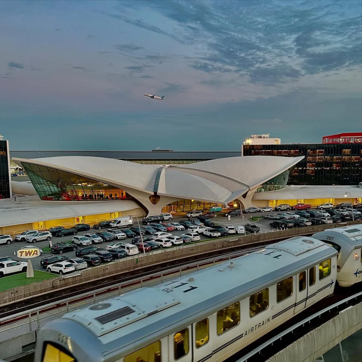 Pemandangan senja ditinggikan dari Pusat Penerbangan TWA patung di New York City, dengan cangkang betonnya yang luas membentuk sayap yang menyapu ke atas seperti burung yang sedang lepas landas, diterangi dari dalam, dengan mobil AirTrain dan tempat parkir di dekatnya.