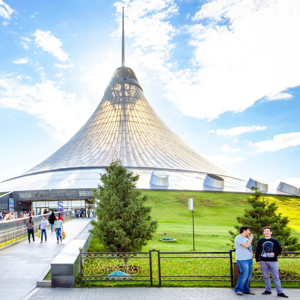 Pemandangan tingkat jalan dari Pusat Hiburan Khan Shatyr berbentuk kerucut transparan yang besar di Astana, menampilkan membran ETFE bercahaya yang didukung oleh tiang tengah masif, di bawah langit biru cerah dengan awan tipis.