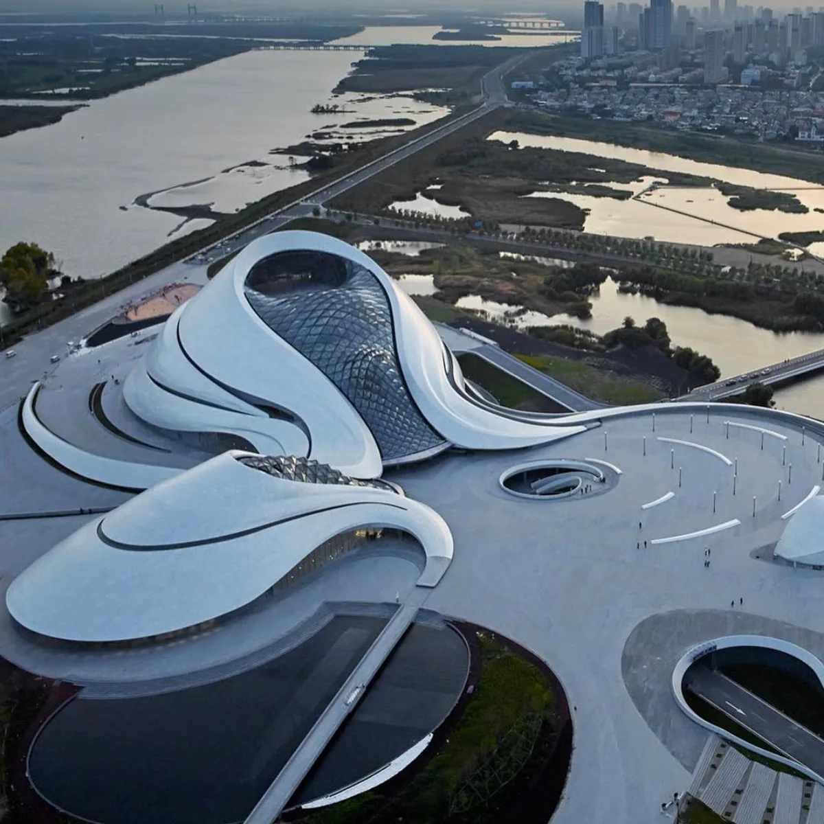 tampilan udara Harbin Opera House yang berbentuk organik putih dengan panel aluminium, dikelilingi lanskap air saat senja