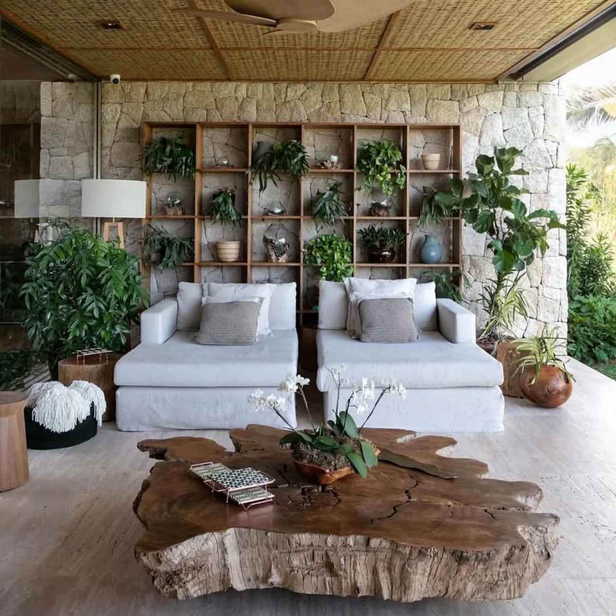 Area santai semi-outdoor gaya Tropis natural dengan plafon anyaman bambu, dinding batu alam, furnitur kayu solid, dan tanaman hias hijau.