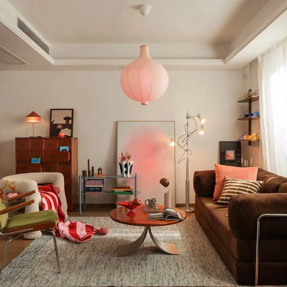 Desain interior ruang keluarga bernuansa Vintage Retro modern, dilengkapi sofa korduroi cokelat, lampu gantung kertas merah muda, dan aksesoris mid-century yang artistik.