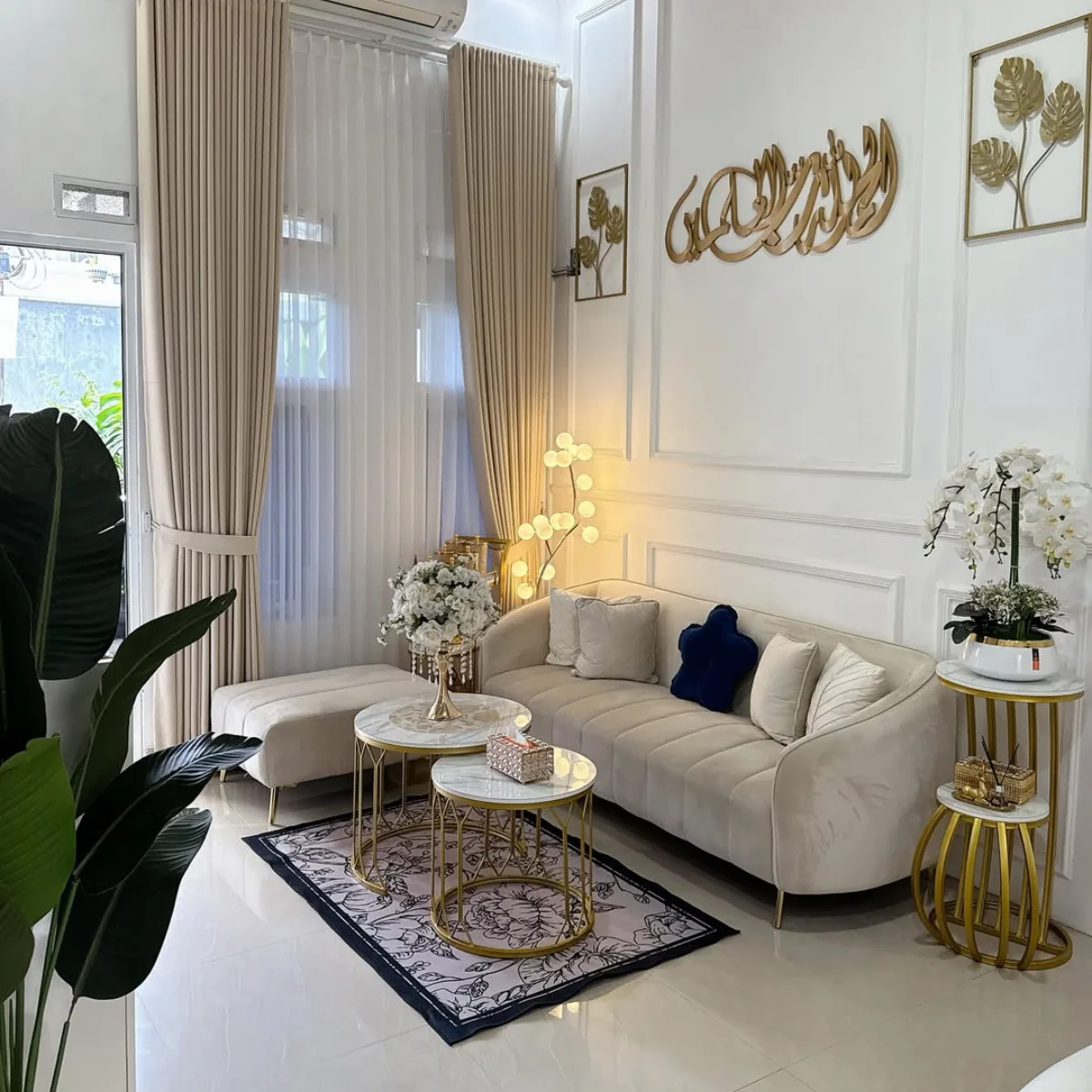 Ruang tamu gaya Modern elegan dengan hiasan dinding kaligrafi emas, meja kopi marmer, gorden tinggi, dan sofa beige yang nyaman.