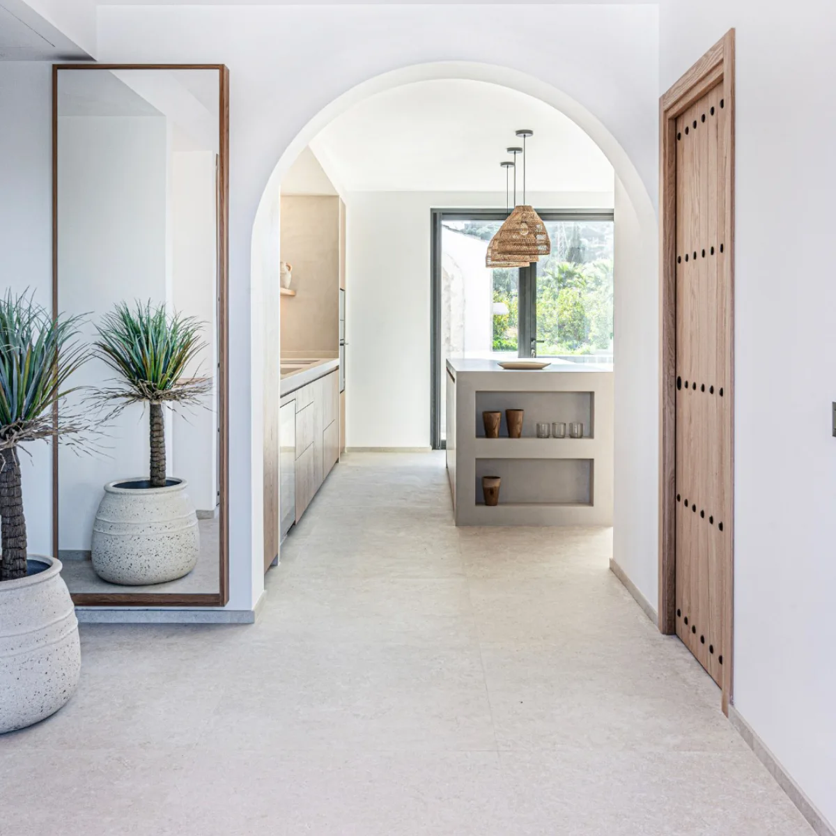 Interior rumah gaya Mediterania dengan lorong pintu lengkung (archway), dinding putih bertekstur, elemen kayu rustic, dan pot gerabah besar.