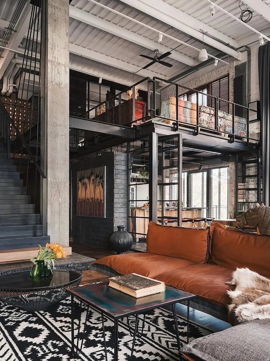 Ruang keluarga berkonsep loft industrial dengan pilar beton, struktur mezzanine besi hitam, dan sofa kulit cokelat