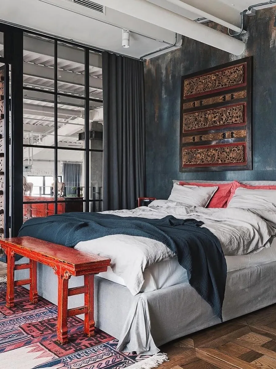 Kamar tidur industrial dengan dinding bertekstur gelap, panel ukiran kayu dekoratif, ranjang abu-abu, bangku kayu merah vintage, dan partisi ruangan dari kaca berbingkai hitam