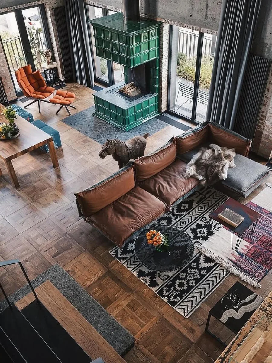 Pemandangan dari atas ruang keluarga industrial yang menampilkan sofa kulit cokelat, karpet bermotif, dan tungku perapian keramik hijau