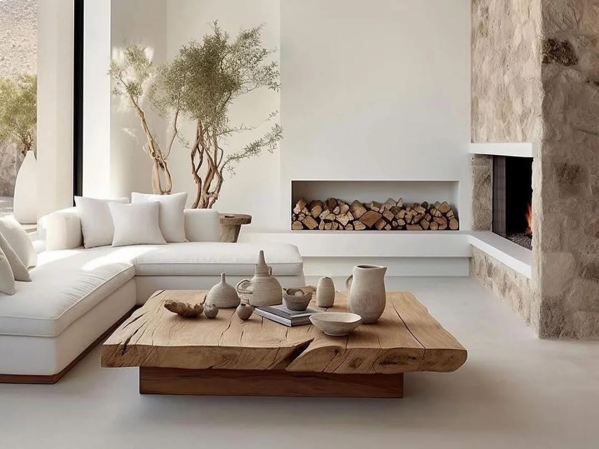 Ruang tamu minimalis bergaya Japandi dengan sofa sudut putih, meja tamu dari potongan kayu solid bertekstur alami, dekorasi vas keramik, dan perapian dengan dinding batu ekspos.