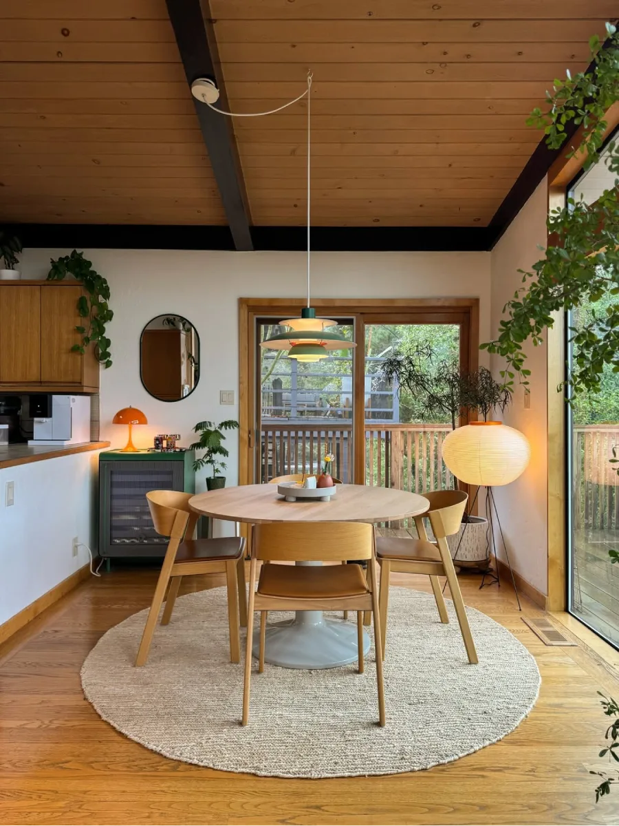 Ruang makan bernuansa Japandi yang hangat dengan meja bundar dan kursi kayu, lampu lantai lampion kertas bergaya Jepang, kabinet hijau, serta pintu kaca geser yang menghadap ke luar.
