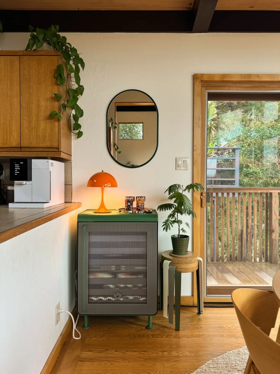 Sudut ruangan estetik menampilkan kabinet penyimpanan beralur warna hijau dengan lampu meja berbentuk jamur warna oranye, cermin dinding oval, dan tanaman hias indoor.
