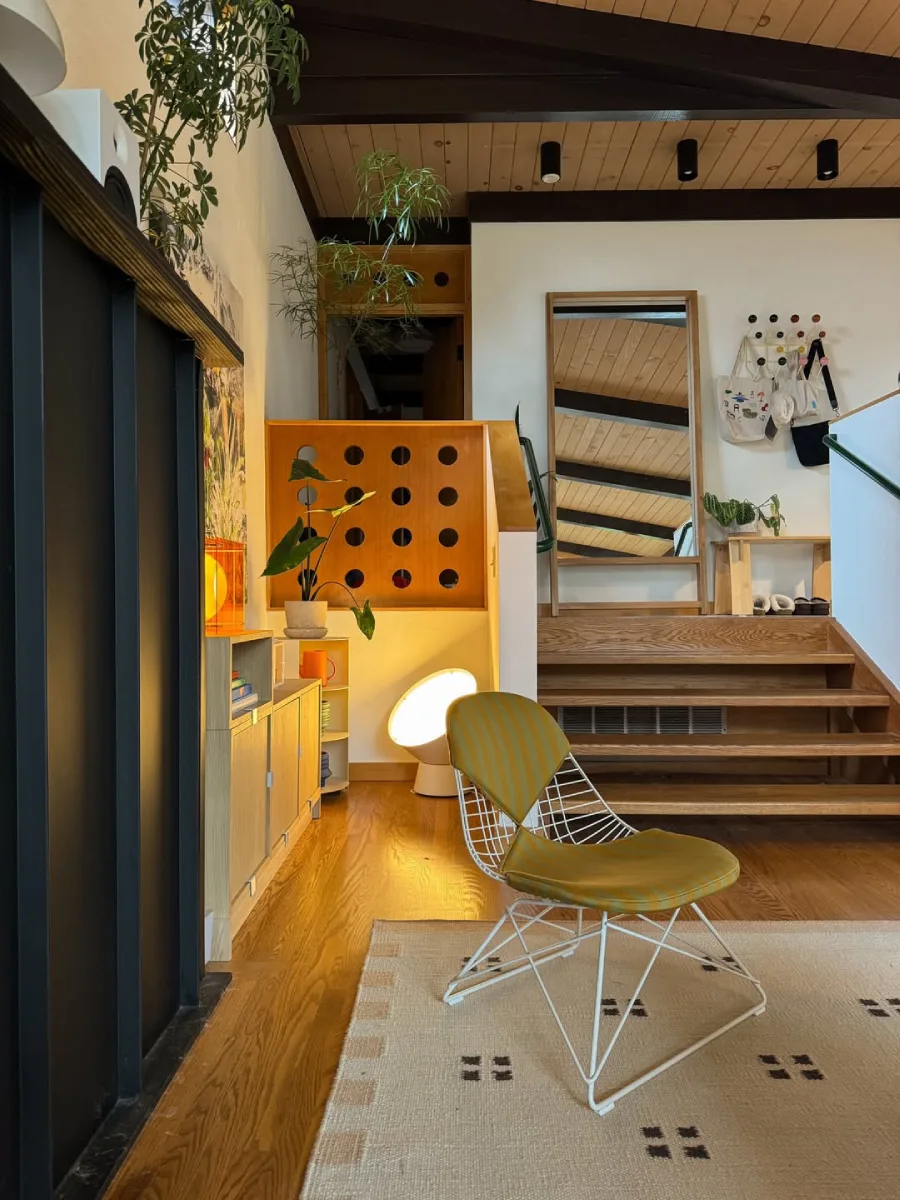 Area sekitar tangga kayu dengan kursi santai berkerangka besi, partisi kayu dengan aksen lubang bundar, cermin lantai, dan pencahayaan hangat dari lampu sudut.