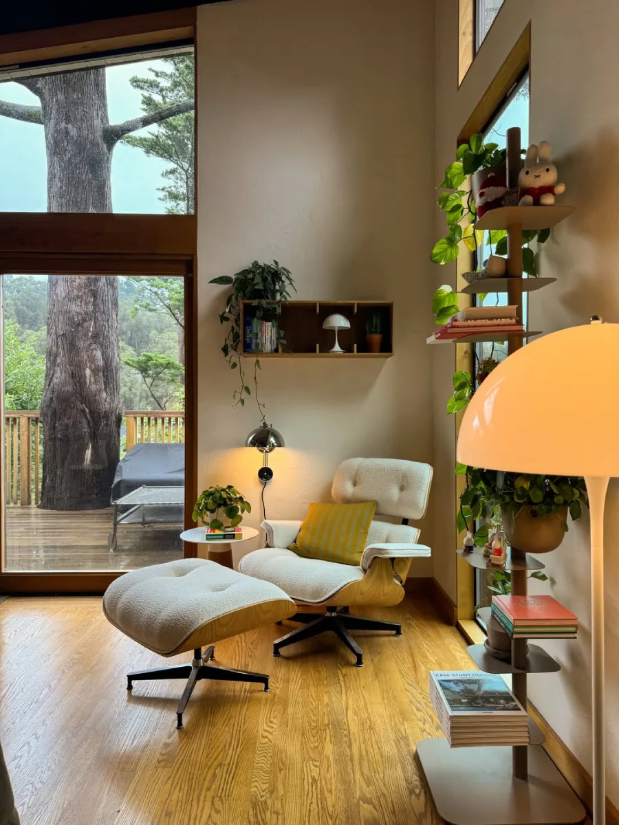 Sudut baca yang nyaman (reading nook) di dekat jendela besar dengan kursi lounge putih, lampu baca, dan rak pajangan vertikal yang dipenuhi tanaman hias serta buku dinding.