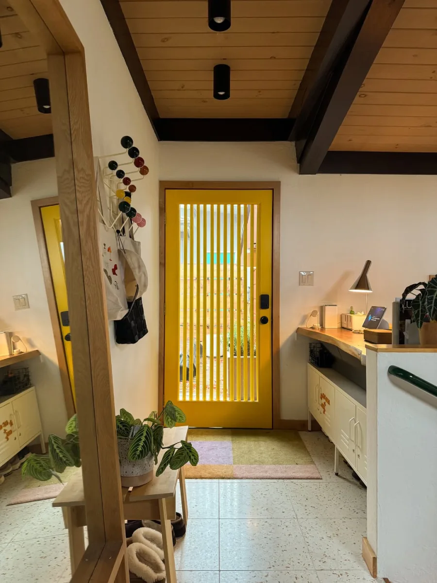 Area pintu masuk (entryway) dengan pintu kuning cerah beraksen garis vertikal, meja konsol kayu, tanaman hias, dan karpet lantai dengan pola blok warna.
