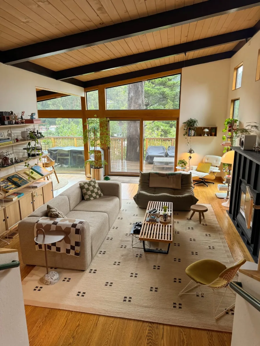 Ruang keluarga luas yang hangat dengan langit-langit kayu berbalok hitam, jendela kaca besar dengan pemandangan alam, perpaduan sofa modern dan kursi lantai, serta karpet bermotif minimalis.