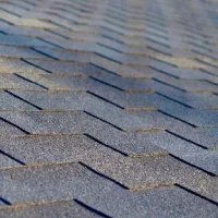 Close-up tekstur atap aspal shingles berlapis dengan pola bertumpuk berwarna abu-abu pada permukaan rumah.