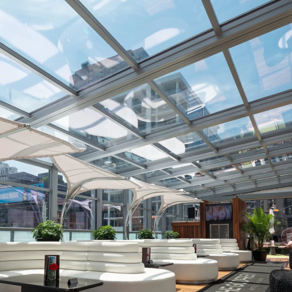 Struktur atap kaca transparan berangka metal yang memberikan pencahayaan alami pada area lounge tempat duduk bersofa putih