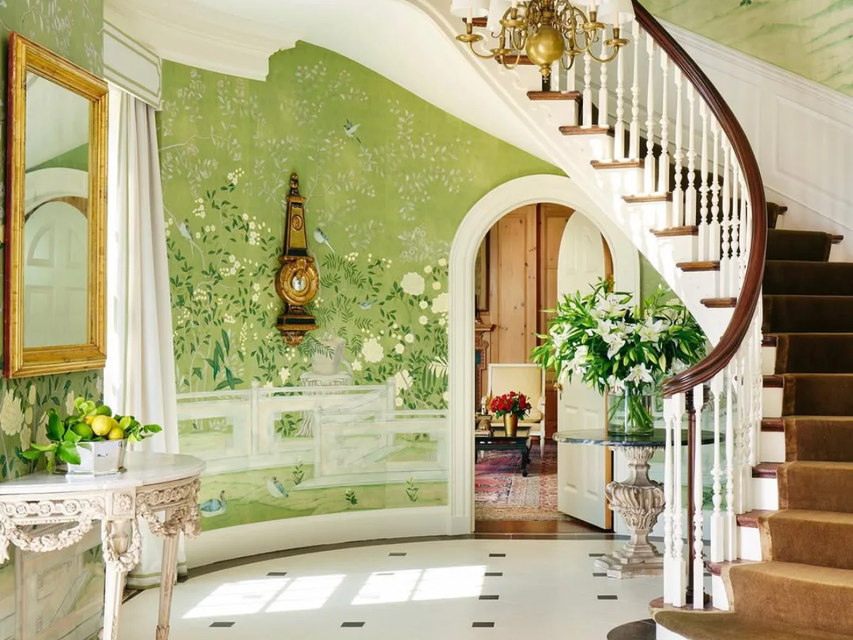 Interior rumah elegan dengan dinding hijau bermotif klasik, tangga melengkung, meja dekoratif, dan bunga segar di ruang masuk yang terang