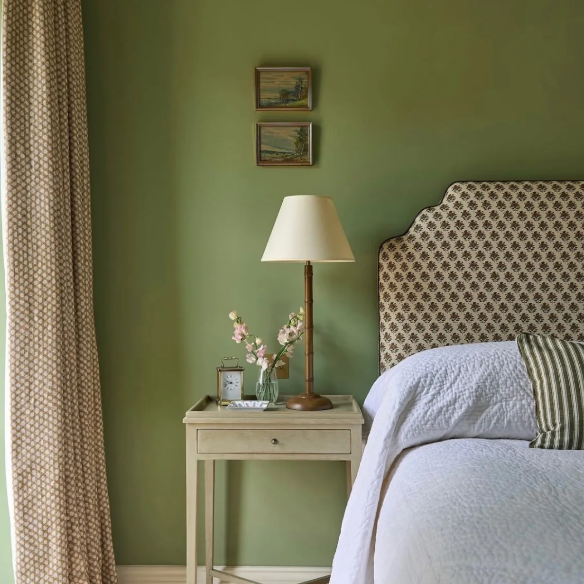 Kamar tidur dengan dinding hijau zaitun lembut, sandaran kepala tempat tidur bermotif geometris, tempat tidur dengan seprai putih dan bantal bermotif garis, meja samping tempat tidur krem dengan lampu rotan, vas bunga, dan jam weker kuningan kecil, serta gorden bermotif di samping jendela dan dua bingkai foto lanskap kecil.