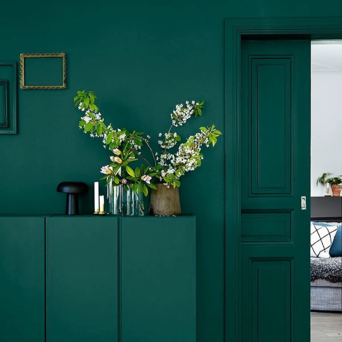 Bufet dan pintu sliding klasik berwarna teal yang serasi dengan dinding, dengan vas berisi cabang bunga sakura, lampu hitam, lilin dalam tempat lilin emas, dan dua bingkai foto emas kosong di dinding, terbuka ke kamar tidur dengan tempat tidur berselimut abu-abu.