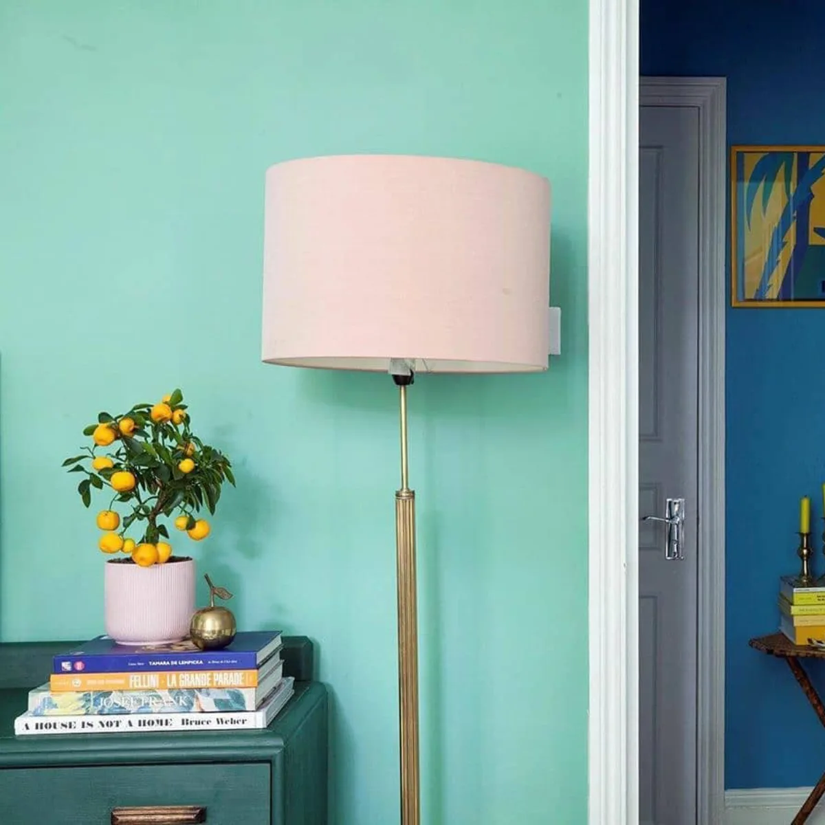 Sudut ruangan dengan meja samping laci hijau tua, tumpukan buku, pot tanaman jeruk mandarin kecil, dan pajangan apel emas, di samping lampu lantai kuningan dengan kap lampu merah muda, dengan latar belakang dinding hijau mint dan pintu terbuka ke ruangan lain dengan dinding biru tua dan meja anyaman.