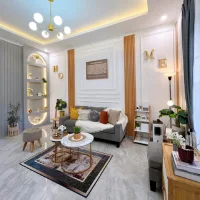 Ruang tamu modern elegan dengan sofa abu-abu, dinding putih beraksen panel molding, rak melengkung dengan lampu hangat, dan dekorasi huruf emas bertuliskan HOME.