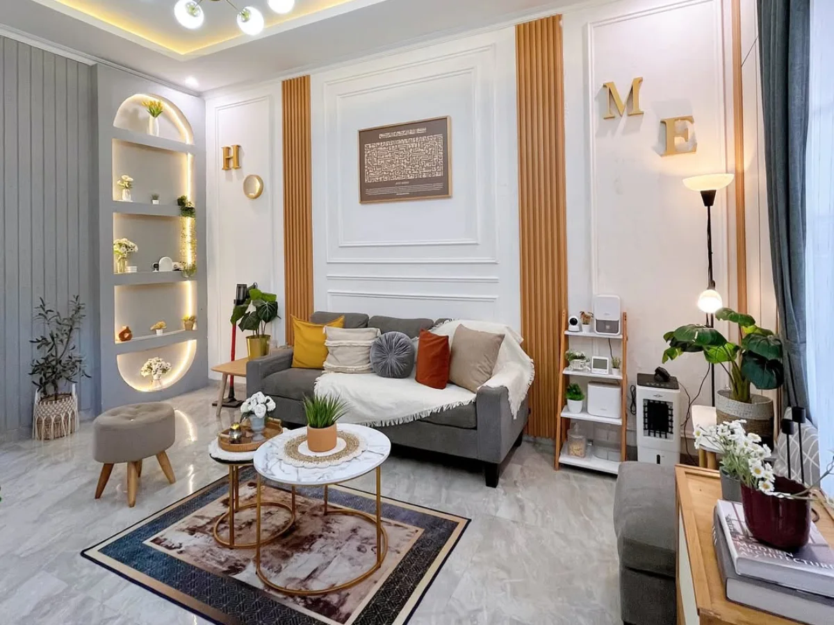 Ruang tamu modern elegan dengan sofa abu-abu, dinding putih beraksen panel molding, rak melengkung dengan lampu hangat, dan dekorasi huruf emas bertuliskan HOME.