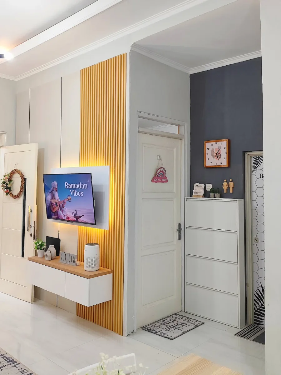 Area hiburan ruang tamu minimalis dengan panel kayu vertikal, rak TV gantung putih, dan kabinet sepatu putih bersusun di sebelah pintu masuk.