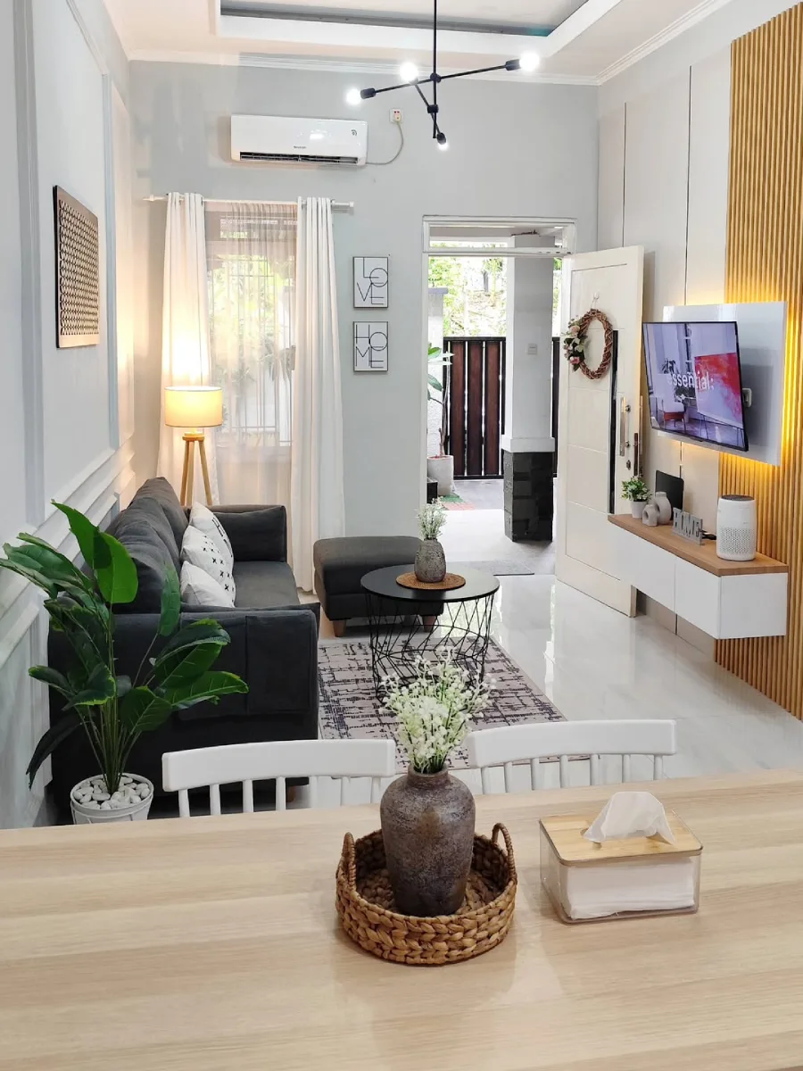 Ruang tamu berdesain minimalis dengan sofa abu-abu, meja kopi hitam bundar, dan meja makan kayu yang menghadap ke pintu utama yang terbuka.