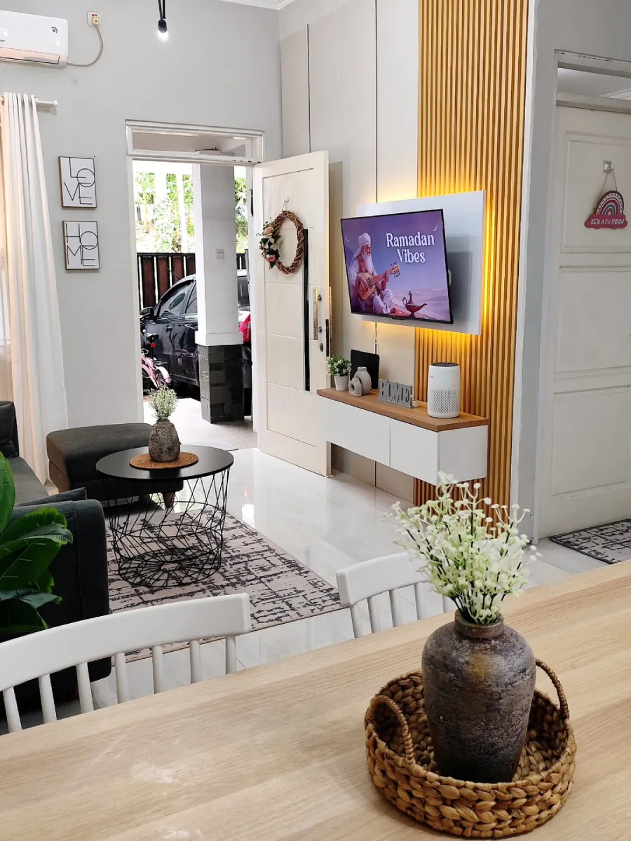 Sudut ruang tamu minimalis yang menampilkan rak TV gantung dengan pencahayaan hangat di belakangnya, sofa abu-abu, dan pintu depan yang terbuka.