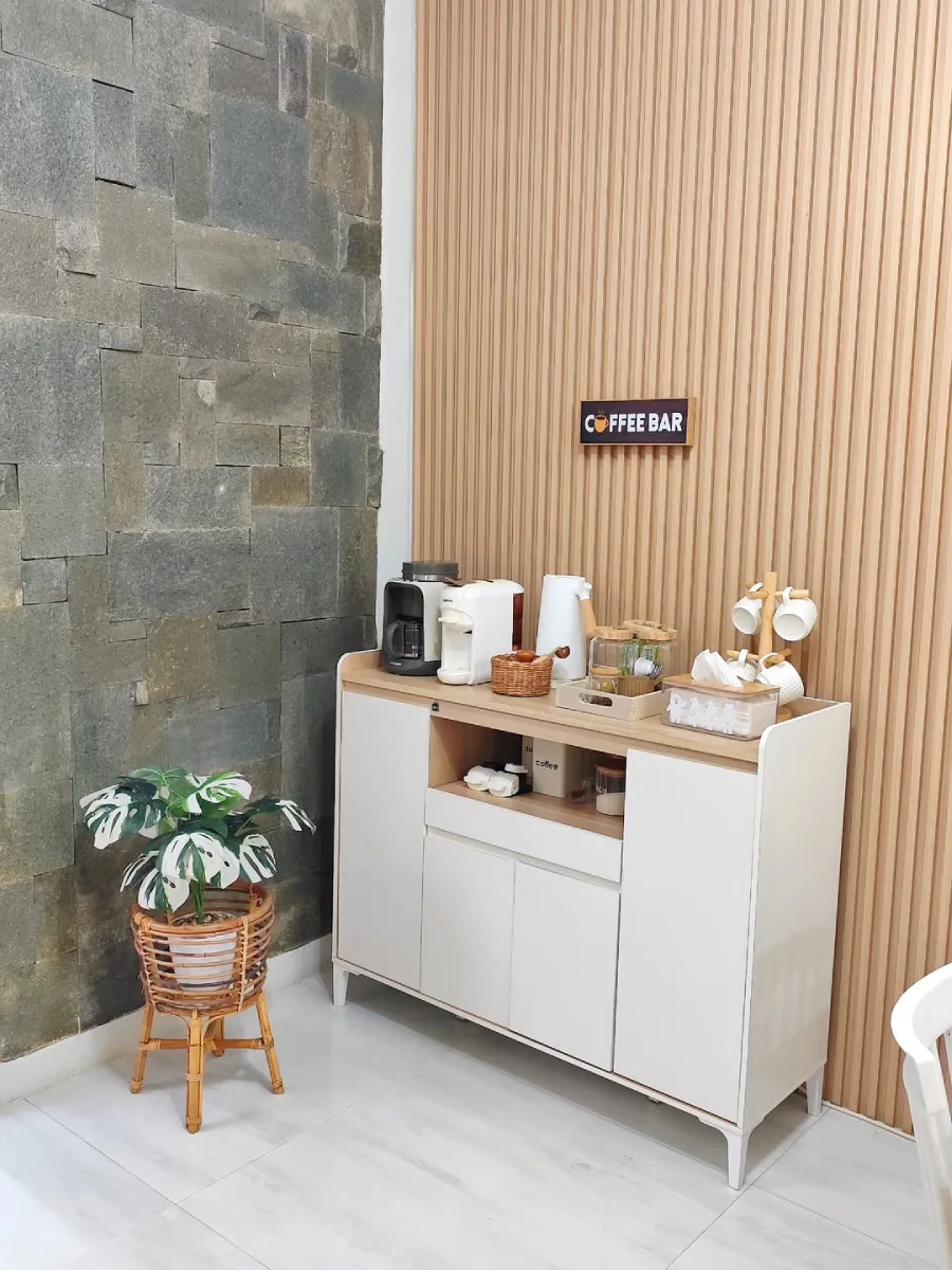 Area sudut kopi minimalis dengan kabinet putih, mesin pembuat kopi, dinding panel kayu, dan tanaman hias monstera di dalam pot anyaman.