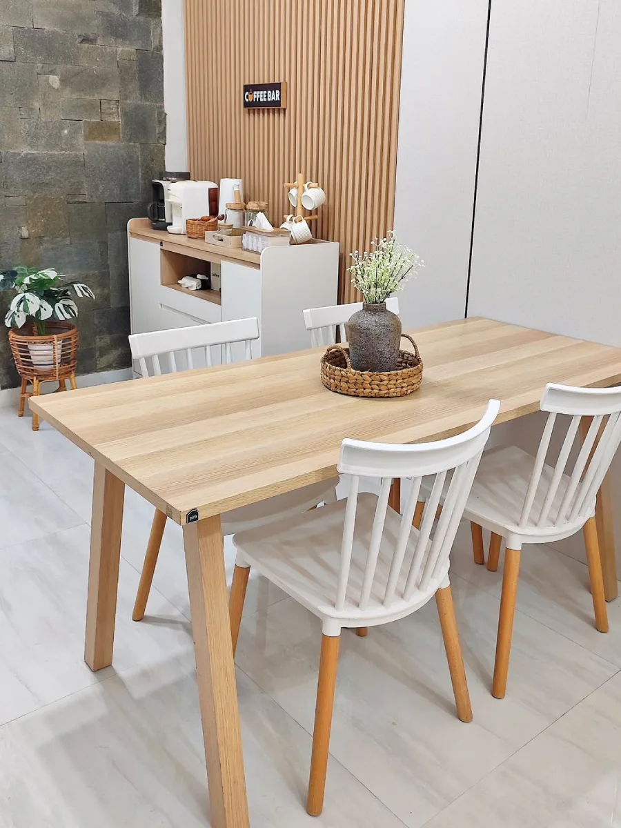 Meja makan kayu bernuansa terang dengan kursi putih bergaya minimalis, dihiasi vas bunga estetik di atas nampan anyaman.