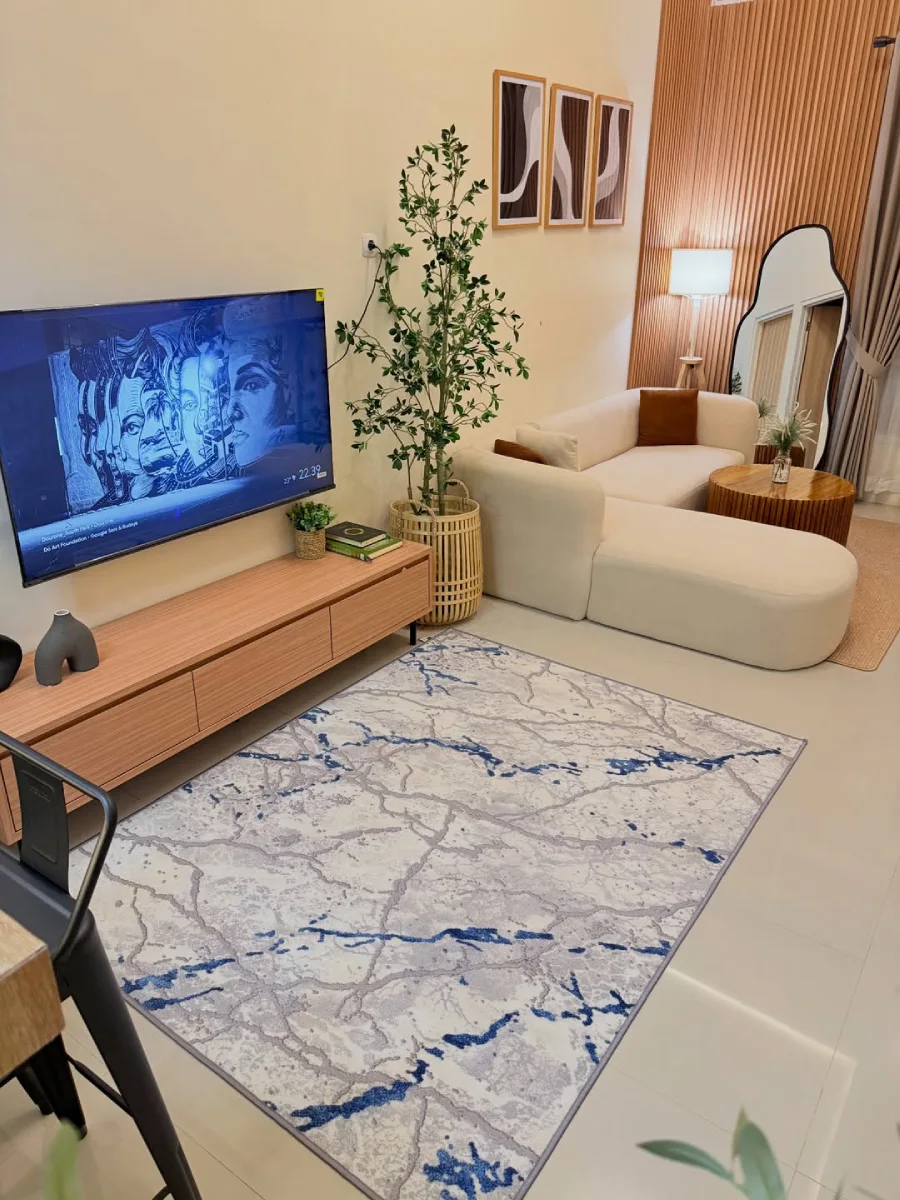 Area menonton TV bergaya minimalis dengan kabinet TV kayu terang, karpet bermotif marmer abu-abu biru, dan tanaman hias artificial di sudut ruangan.