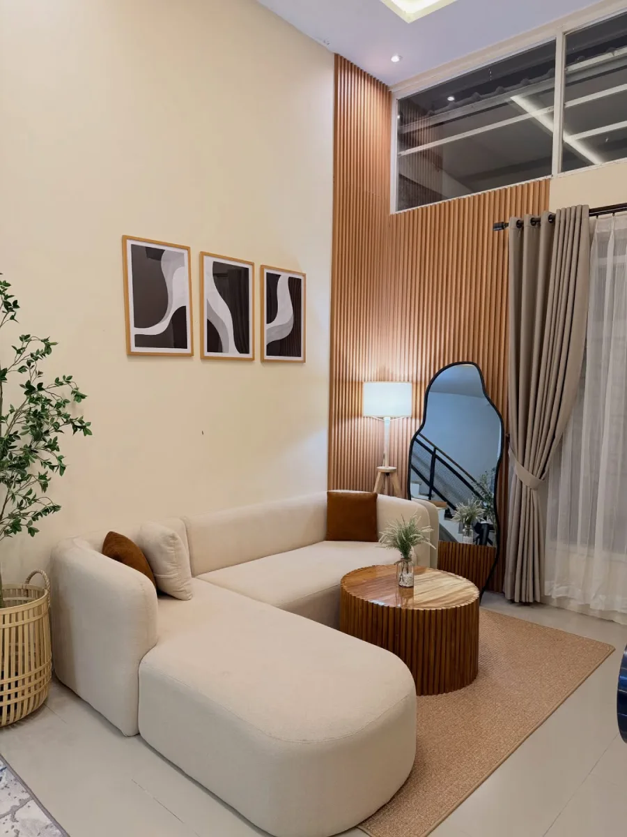 Sudut santai di ruang tamu bersiling tinggi dengan sofa L krem, tiga pajangan seni abstrak monokrom, dan cermin estetis bergelombang.