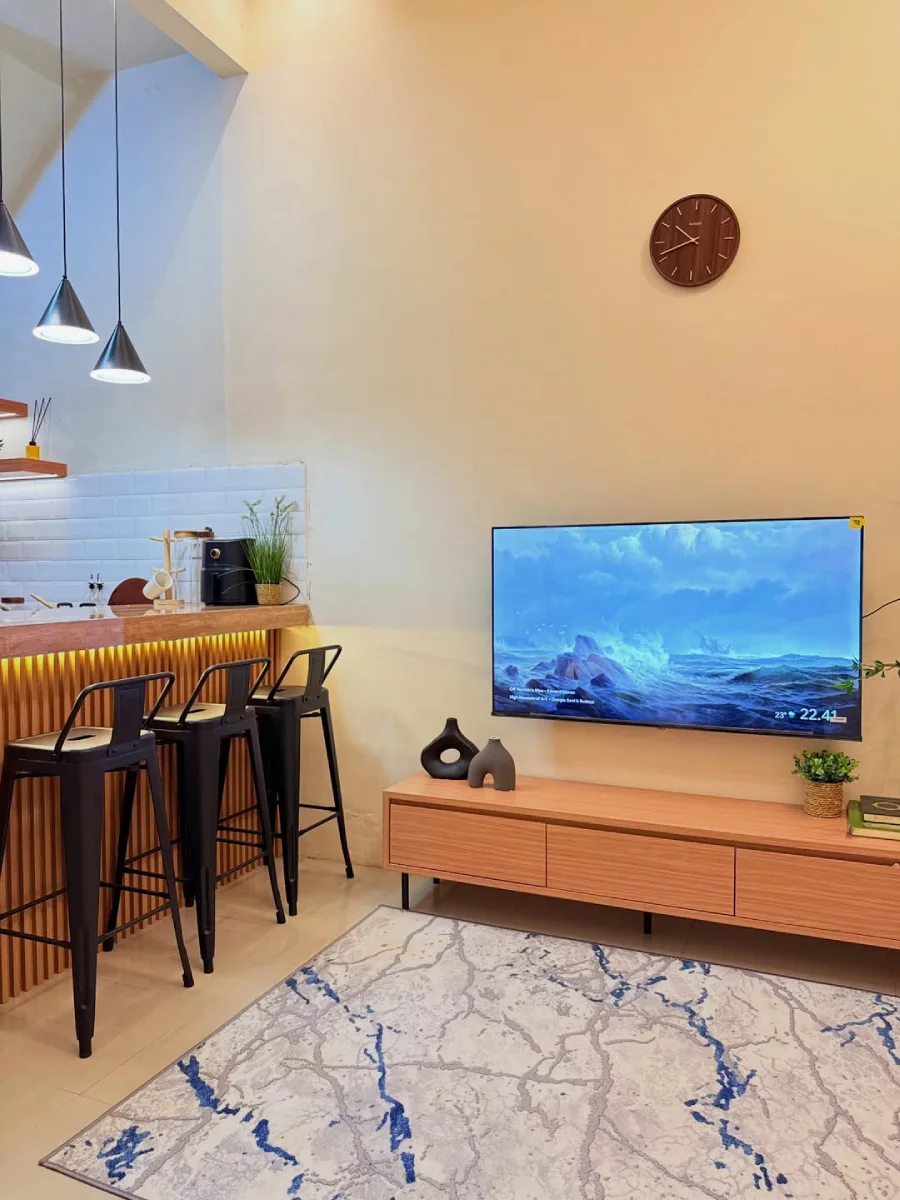 Transisi ruang keluarga ke dapur minimalis yang menampilkan TV layar datar di dinding, kabinet kayu, dan meja bar beraksen kayu dengan tiga kursi barstool hitam.