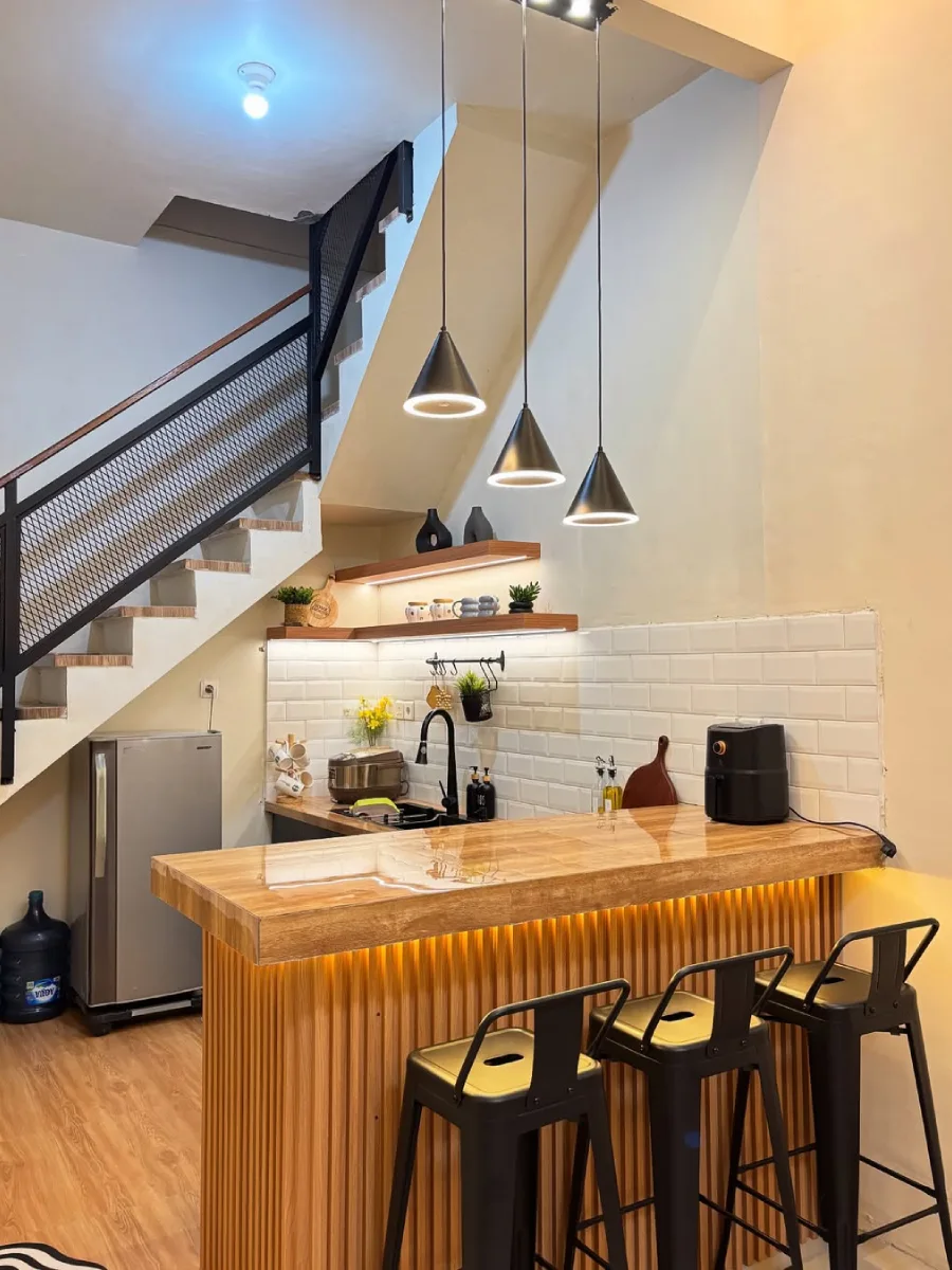 Pemanfaatan ruang cerdas untuk dapur minimalis di bawah tangga, dilengkapi meja peninsula berlapis kayu, kursi bar hitam, dan tiga lampu gantung modern.