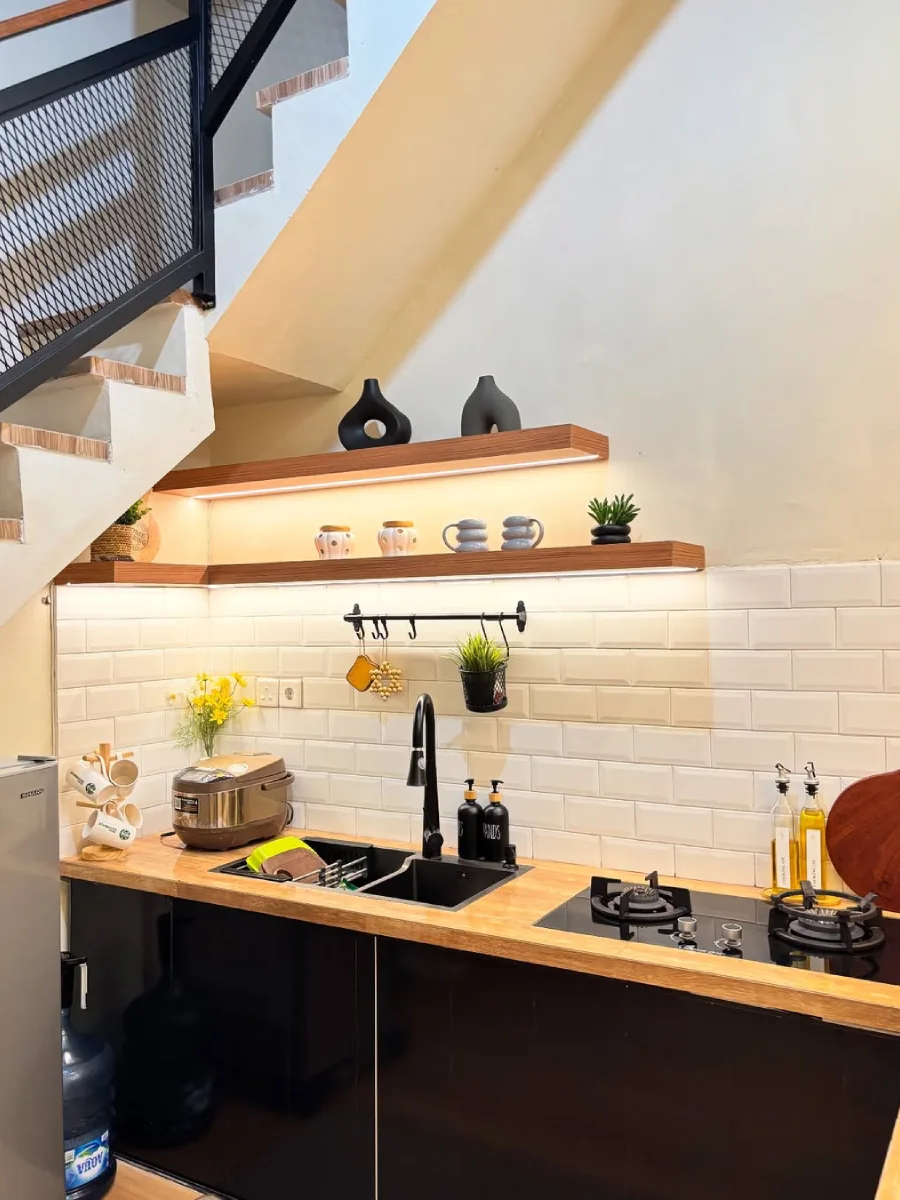 Detail area kitchen set di bawah tangga dengan meja dapur butcher block, backsplash keramik subway tile putih, rak ambalan kayu, dan kompor tanam.
