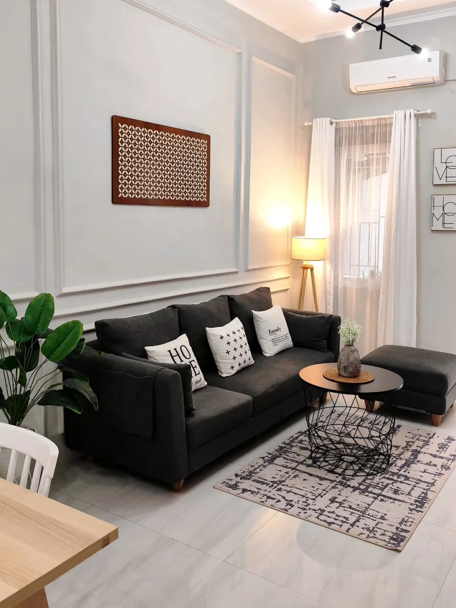 Ruang keluarga bergaya minimalis dengan sofa abu-abu gelap, bantal dekoratif monokrom, meja kopi rangka besi, dan hiasan dinding ukiran kayu pada dinding berpanel abu-abu muda.