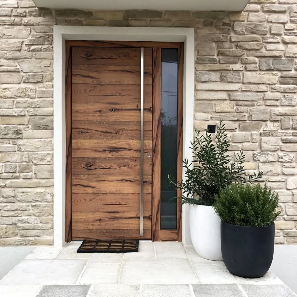 Pintu kayu solid tekstur alami dengan handle stainless steel vertikal dan dinding batu alam.