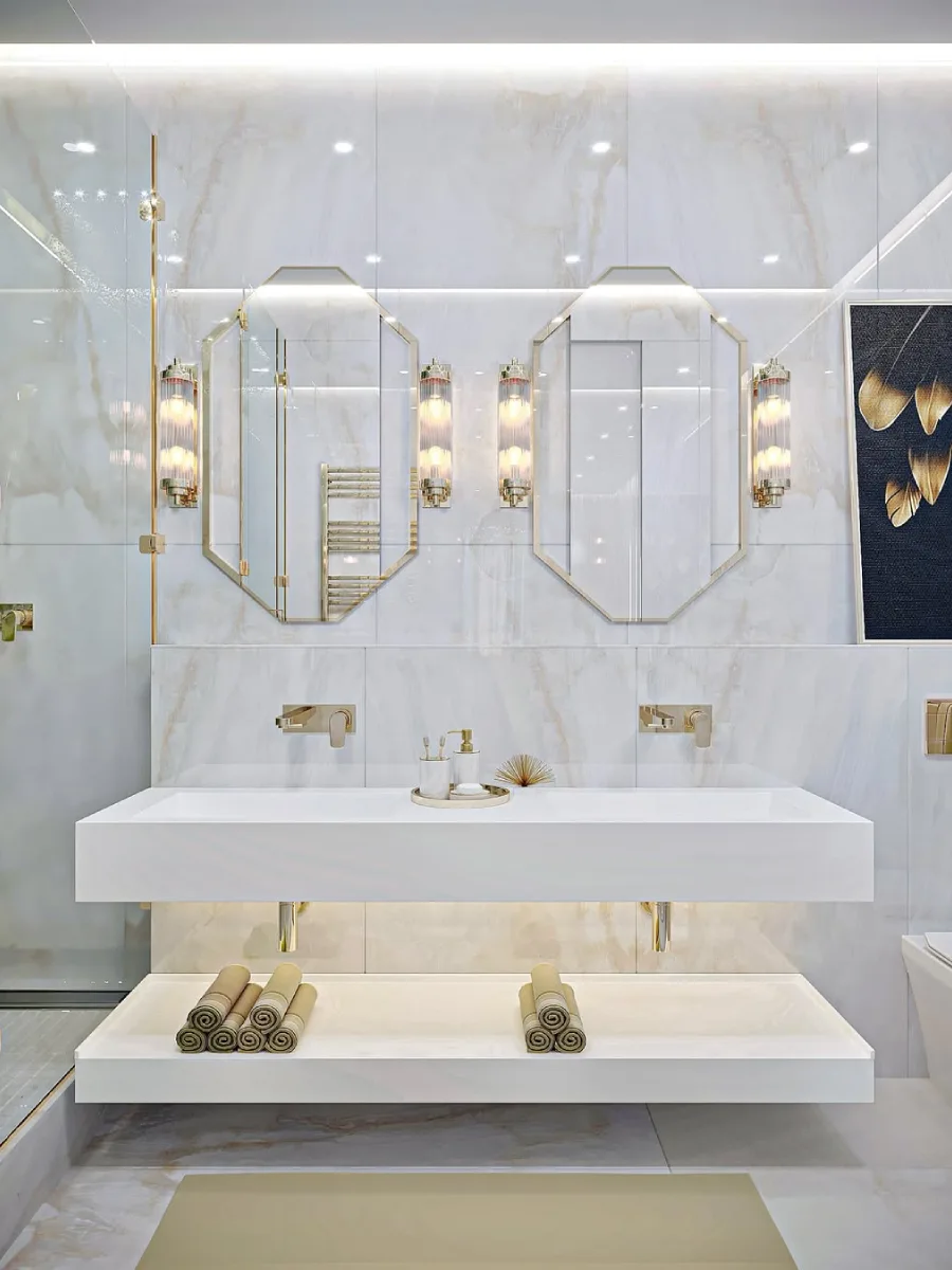 Wastafel kamar mandi ganda mengambang dengan cermin segi delapan, keran emas, sconce dinding tabung kaca, dan ubin marmer putih.