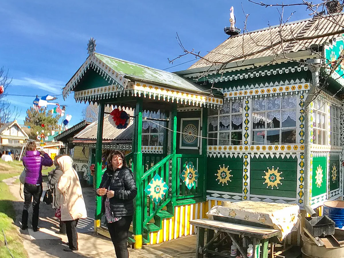 Rumah kayu tradisional Rusia berwarna hijau dengan ukiran dekoratif khas, dilengkapi teras kecil, terlihat beberapa orang berdiri di depan rumah pada hari cerah.
