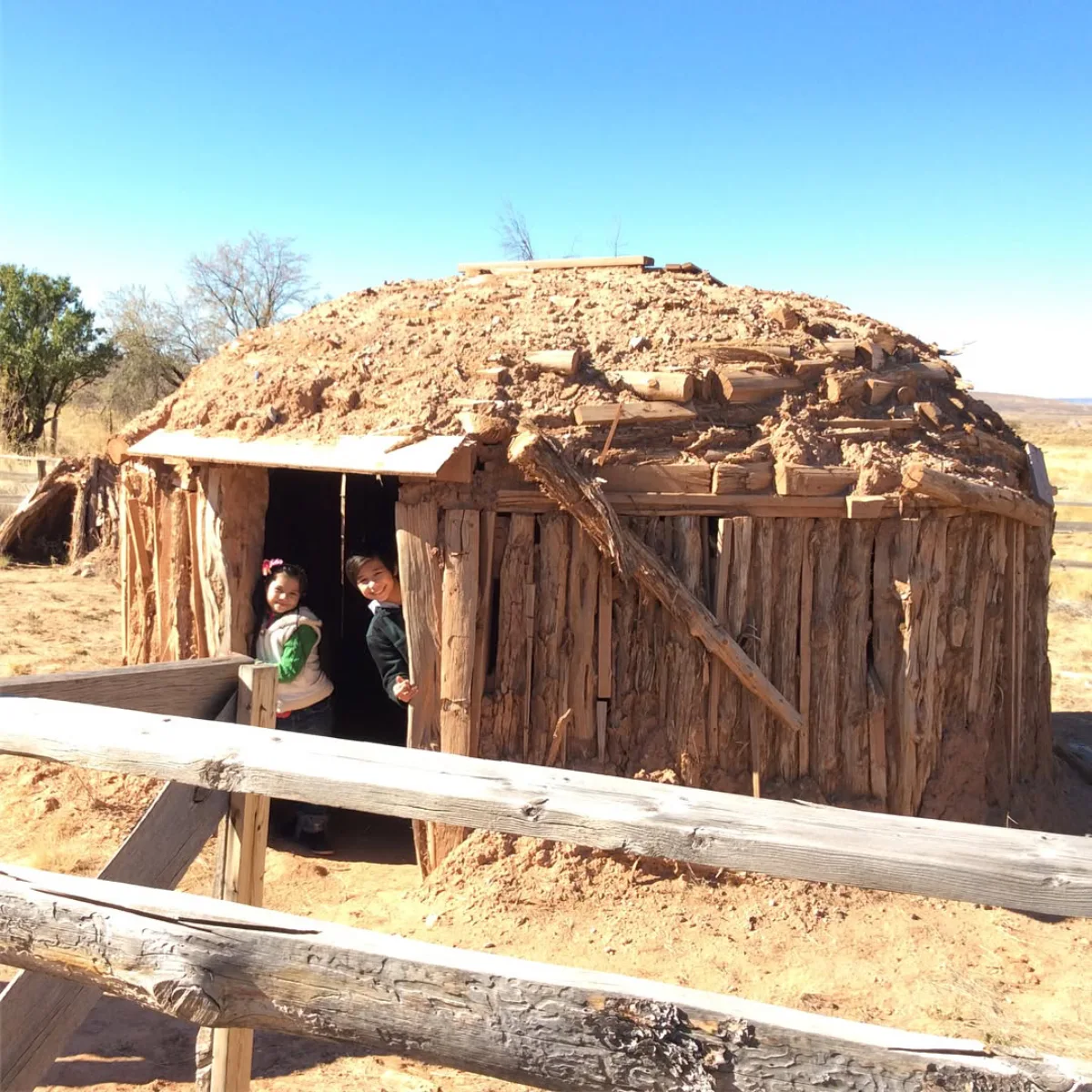 Dua anak kecil mengintip dari pintu masuk sebuah hogan tradisional Navajo yang terbuat dari kayu dan tanah di bawah langit biru yang cerah.