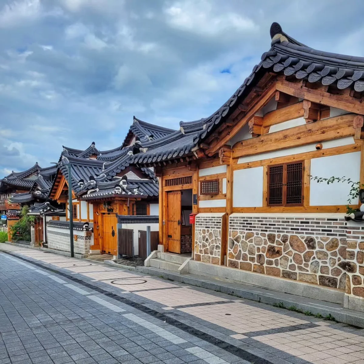 Pemandangan jalan Hanok tradisional Korea di desa Bukchon. Sederetan rumah Hanok dengan atap genteng keramik gelap yang melengkung dan dinding kayu-putih berbaris di sepanjang jalan beraspal. Pintu masuk kayu dan jendela kisi-isi terlihat di bawah langit mendung kebiruan.