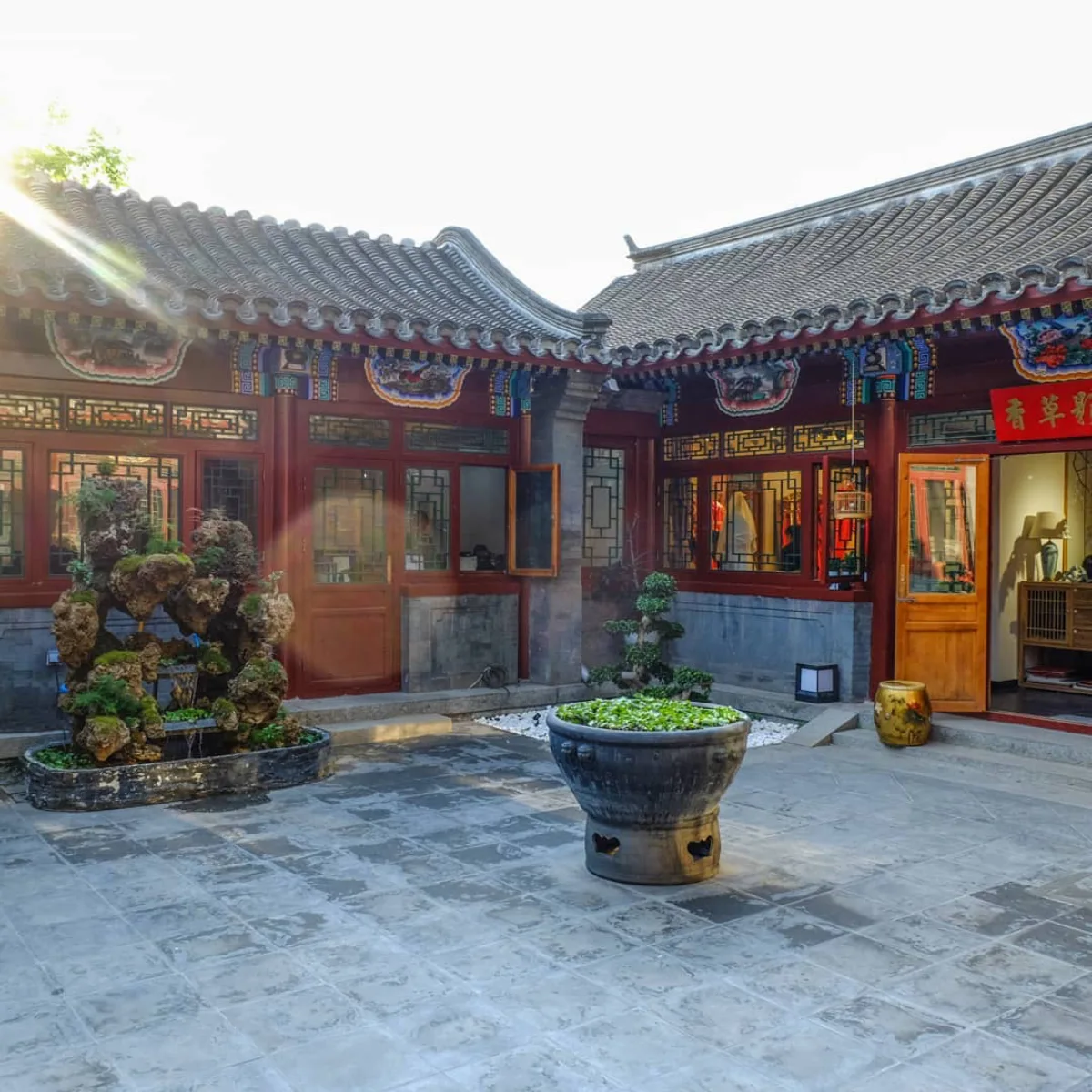 courtyard tengah yang luas dari sebuah kompleks Siheyuan Tiongkok kuno. Bangunan dengan atap genteng tradisional mengelilingi halaman, menampilkan tiang-tiang kayu merah dan ukiran rumit. Terdapat hiasan batu di kiri dan pot tanaman besar di tengah, dengan jalur batu dan langit cerah.