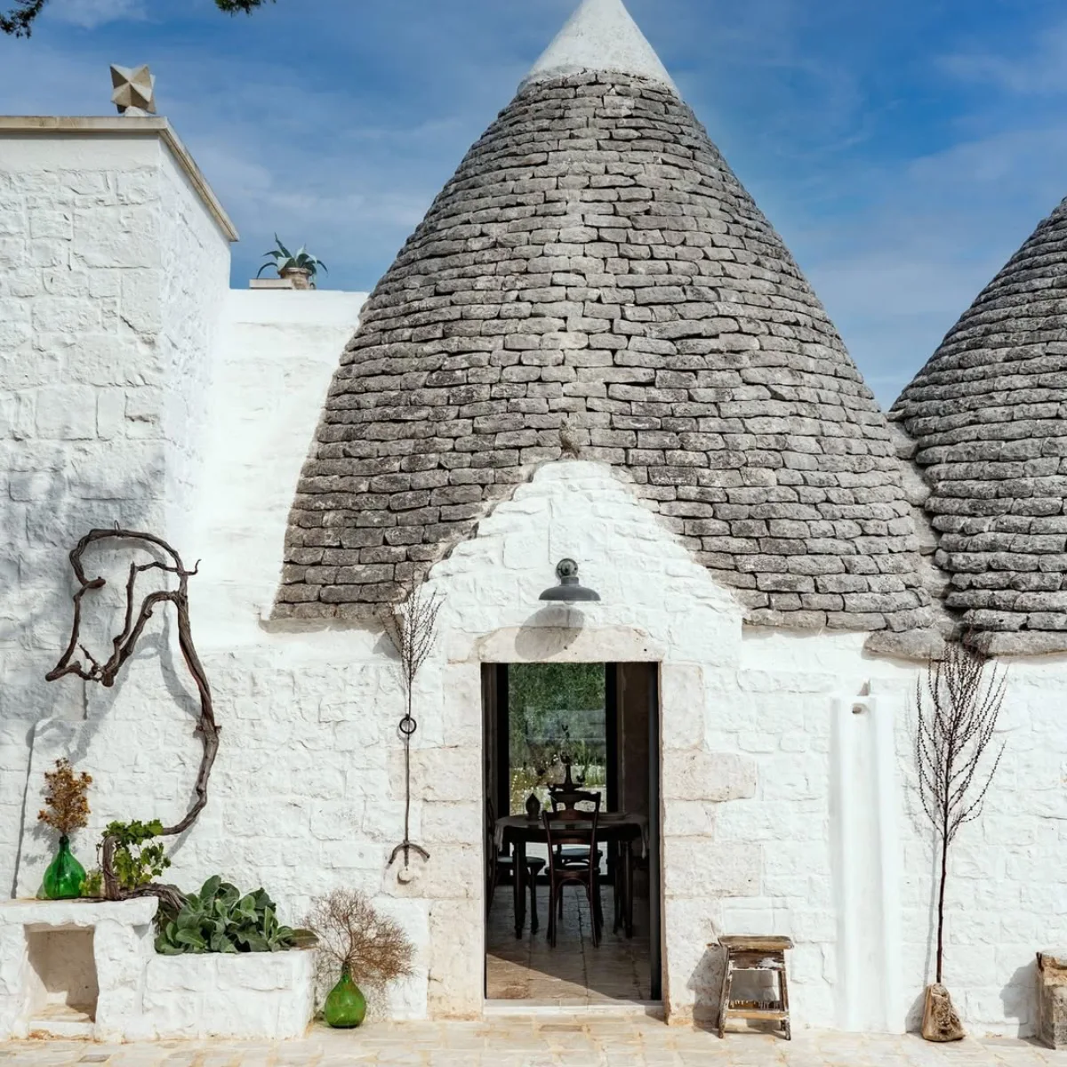 Sebuah foto dekat dari fasad trullo batu putih tradisional. Atap kerucut yang khas terbuat dari batu abu-abu tumpuk, dengan dekorasi puncak batu putih. Pintu masuk mengarah ke ruang makan dengan meja dan kursi, dengan tanaman di rak batu di luar.
