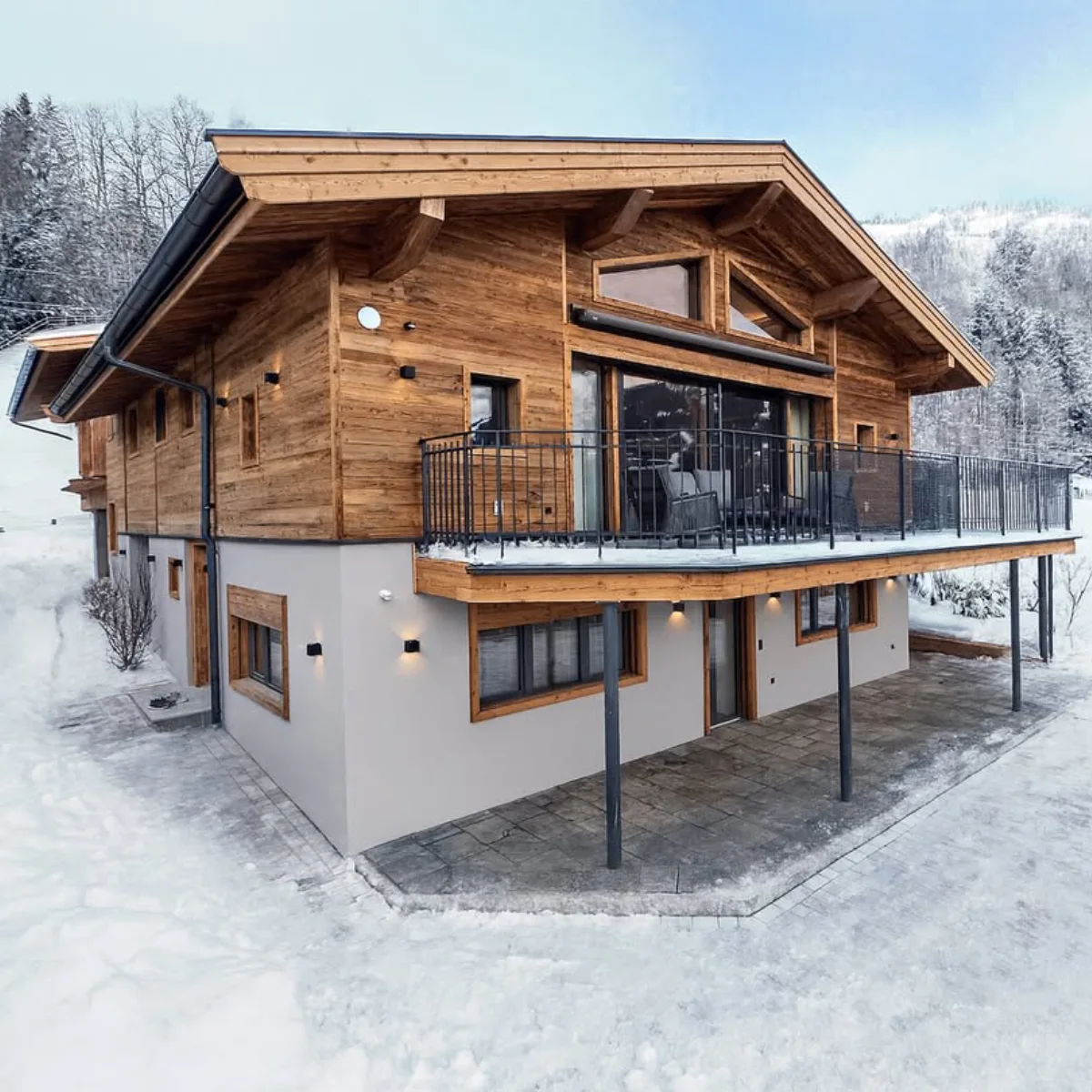 Sebuah foto miring dari sebuah Chalet Swiss modern-klasik berbahan kayu dengan balkon besi tempa yang luas. Lantai dasarnya adalah beton abu-abu, dan atap kayunya menjorok ke luar. Rumah ini dikelilingi oleh salju tebal dan pepohonan pinus di lereng gunung.